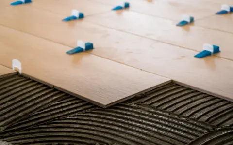 Pose de carrelage 60x60 en cours. Des carreaux en bois clair sont posés sur du mortier frais, avec des croisillons bleus et blancs pour un espacement parfait.