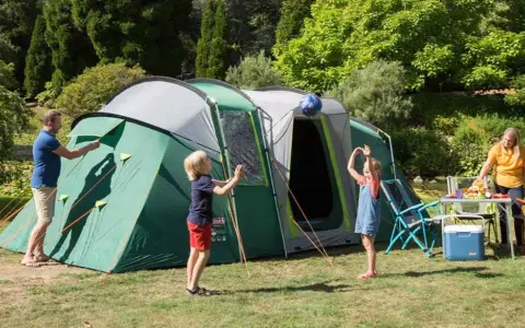 Radosny camping rodzinny: tata rozkłada namiot, dzieci bawią się piłką, mama przygotowuje posiłek.