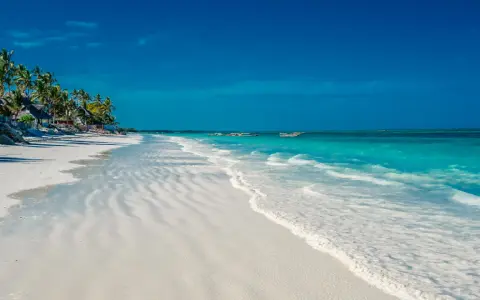 En vit sandstrand med turkost vatten och palmer vid Kendwa, Zanzibar. Båtar guppar i fjärran.