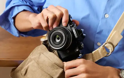 Osoba w niebieskiej koszuli wkłada czarny aparat Nikon z obiektywem do torby.