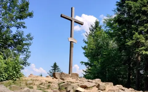 Drewniany krzyż na skalistym szczycie w Górach Świętokrzyskich. Otaczają go bujne drzewa i błękitne niebo z chmurami.