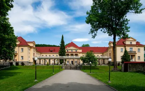 Eleganckie, żółte budynki z czerwonymi dachami, przypominające stare łazienki Krynica Zdrój, otoczone zielonym ogrodem.