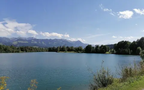 Blauer Himmel über dem Sonthofener See, umgeben von grünen Bäumen und majestätischen Bergen.