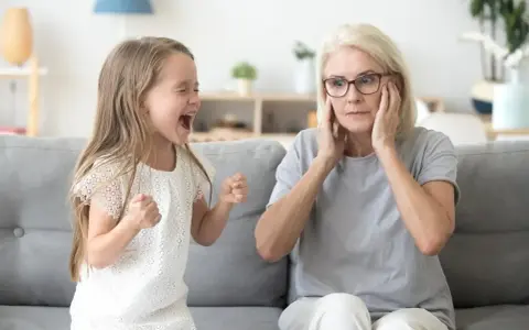 Mała dziewczynka krzyczy, gdy dziecko rządzi rodzicami. Babcia zatyka uszy, wyraźnie zirytowana.