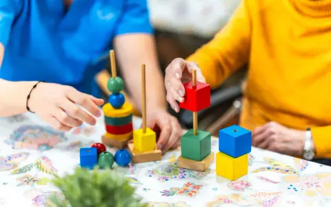Manos de un terapeuta y un adulto mayor jugando con bloques de colores. Un ejemplo de qué es la terapia ocupacional para estimular la motricidad fina.