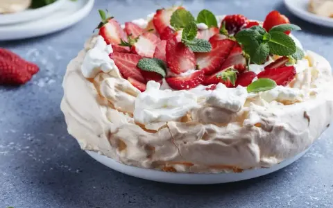 Pyszny tort bezowy z mascarpone i bitą śmietaną, ozdobiony świeżymi truskawkami i miętą.