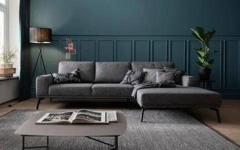 Ein moderner Wohnbereich mit einem anthrazitfarbenen Sofa, das auf einem gemusterten Teppich steht. Ein Couchtisch mit einem offenen Buch und eine Stehlampe vervollständigen das Bild.