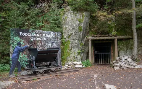 Wejście do Podziemnego Miasta Osówka, z wagonikiem i tablicą informacyjną. Widać skalne ściany i wejście do tunelu.