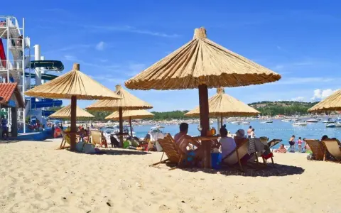 Słoneczna plaża na wyspie Rab, z ludźmi odpoczywającymi pod strzechami, w tle widać wodę z łodziami i zjeżdżalnie wodne.