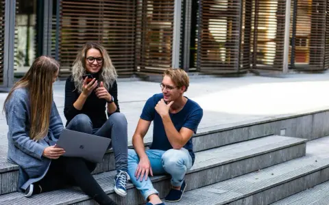 Trójka studentów, w tym jedna osoba na studiach lingwistycznych, siedzi na schodach, korzystając z laptopa i telefonu.