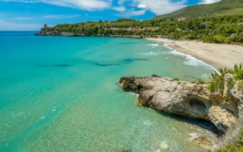 Spiaggia dorata e mare cristallino nel Cilento, con una torre antica su una scogliera e colline verdi sullo sfondo.