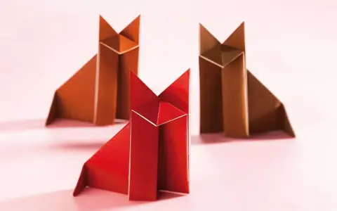 Drei origami Füchse, einer rot, zwei braun, sitzen auf rosa Untergrund.
