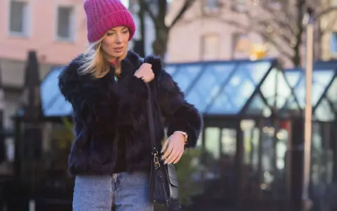 Krótkie futerko stylizacje: blondynka w różowej czapce i czarnym futerku, z jeansami i torebką.