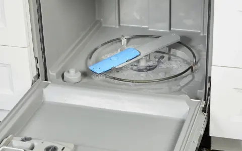 Water and suds fill the bottom of a dishwasher, with a blue spray arm component visible. This image shows a step in how to fix a clogged dishwasher.