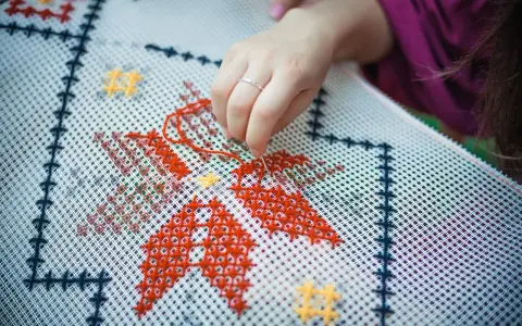 Dłoń z igłą i czerwoną nitką wyszywa wzór na białej kanwie. Widać fragmenty czerwonych i żółtych gwiazdek. Zamek otwiera się w kierunku ręki.