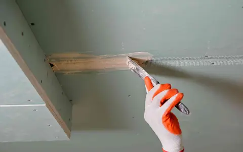 Ręka w rękawicy nakłada masę szpachlową na połączenie płyt gipsowo-kartonowych, przygotowując je do dalszych prac. Klejenie płyt GK na ściany wymaga precyzji.