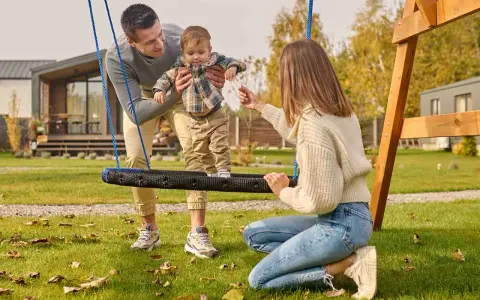 Rodzina cieszy się radosnymi zabawami na polu. Tata trzyma dziecko na huśtawce, a mama wyciąga do niego ręce.