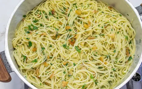 Espaguetis al ajillo recién hechos, con trocitos de ajo dorado y perejil fresco, servidos en una sartén metálica.