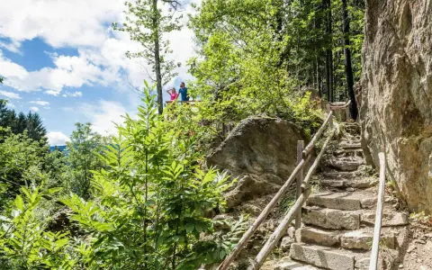 Ein Paar genießt die Aussicht vom Schenner Waalweg. Steintreppen führen durch üppige Vegetation und Felsen.