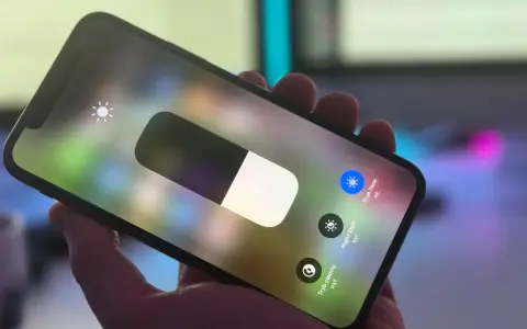 Dłoń trzyma iPhone'a z włączonym panelem sterowania. Widoczny suwak jasności i ikona True Tone.