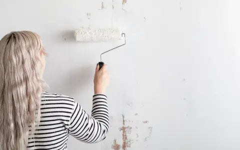 Mujer con cabello rubio y camiseta a rayas está a punto de empapelar pared con rodillo blanco.