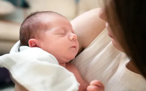 Śpiące niemowlę w ramionach mamy. Warto sprawdzić, ile powinno jeść niemowlę -- tabela, by zapewnić mu najlepszy start.