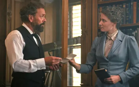 En scen från en alfred nobel film. En man med skägg ger ett brev till en kvinna i en blå dräkt framför en bokhylla.