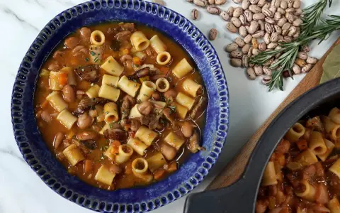 Un reconfortante plato de pasta e fagioli en un cuenco azul, con frijoles y romero esparcidos alrededor.