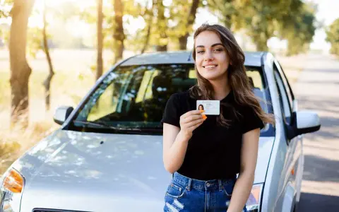 Szczęśliwa młoda kobieta z prawem jazdy od 16 lat stoi obok samochodu na tle drzew.