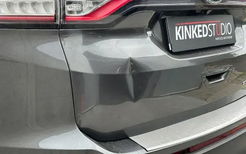 Widoczne wgniecenie w tylnej klapie szarego Forda. Tabliczka "KINKEDSTUDIO" sugeruje miejsce naprawy.