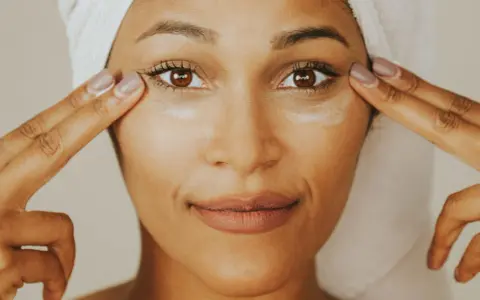Une femme applique de la crème contour des yeux sur tout le visage, avec une serviette sur la tête.