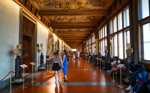 Wnętrze muzeum Uffizi z rzeźbami i malowanym sufitem. Ludzie spacerują i odpoczywają wzdłuż korytarza.