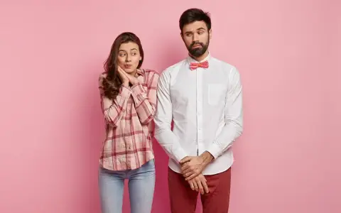 Coppia su sfondo rosa. Lei, con camicia a quadri e jeans, sembra timida. Lui, con camicia bianca e papillon, è pensieroso. Come vestirsi al primo appuntamento uomo?