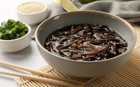 Miseczka z makaronem soba w sosie, posypana sezamem. Obok miseczki z posiekanym szczypiorkiem i sezamem. Zdrowy posiłek, mało makaron soba kcal.