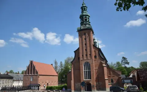 Majestatyczna kolegiata w Kartuzach z cegły, z zieloną wieżą i zegarem, na tle błękitnego nieba z chmurami.