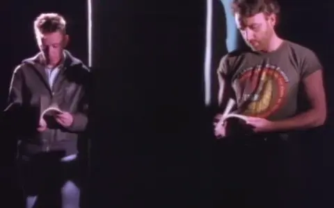 Dwóch mężczyzn czyta. Jeden w kurtce, drugi w koszulce z napisem. Atmosfera jak z "Blue Monday" New Order.