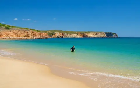 Ein Mann im Neoprenanzug steht im türkisfarbenen Wasser am Praia do Martinhal. Im Hintergrund ragen ockerfarbene Klippen auf.