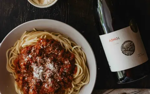 Plato de espaguetis con salsa boloñesa y queso rallado, acompañado de una botella de vino español Adnos.