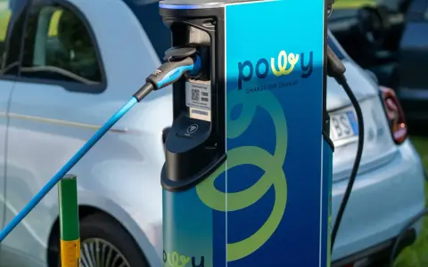 Estación de recarga para coche eléctrico con cable azul conectado. La estación tiene el logo "powy" y el lema "CHARGE THE CHANGE".