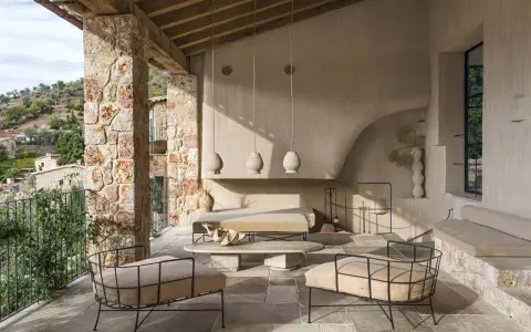 Acogedora terraza interior piso con sillones de metal, sofá y mesa de centro de piedra, bajo un techo de madera y rodeada de muros de piedra.