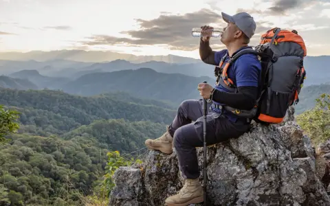Backpacker drinks water on a rocky outcrop, contemplating how much water to bring backpacking for this stunning mountain vista.