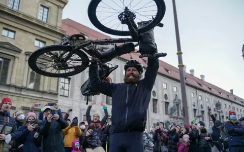 Jonas Deichmann, sichtlich erschöpft, hält sein schlammbedecktes Fahrrad über den Kopf. Eine jubelnde Menge mit Smartphones feiert seine Ankunft.