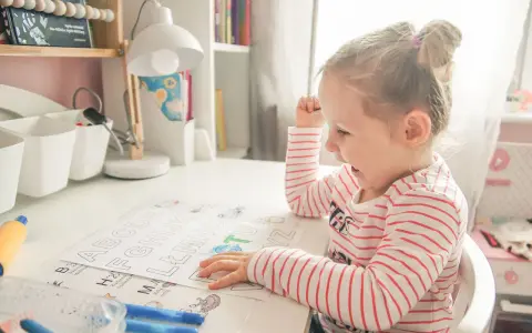 Mała dziewczynka z zapałem uczy się literek dla 3 latka. Na biurku ma kolorowanki z alfabetem i flamastry.