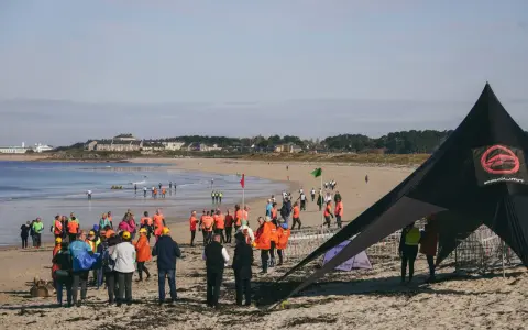 Rassemblés sur la plage pour une course de longe côte, des participants en tenues colorées attendent le départ sous un ciel clair.
