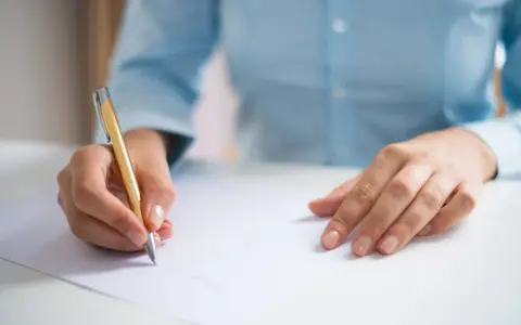 Dłoń pisze złotym długopisem na białej kartce, przygotowując się do egzaminu CAE practice tests.