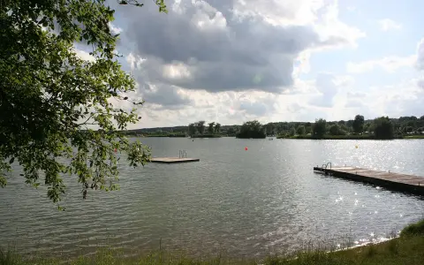 Ein sonniger Tag am Oberrieder Weiher Camping. Ein Steg mit Leiter lädt zum Baden ein, während im Hintergrund Bäume und eine idyllische Landschaft zu sehen sind.