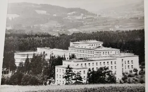 Duży budynek sanatorium w otoczeniu drzew, na tle pagórkowatego krajobrazu.