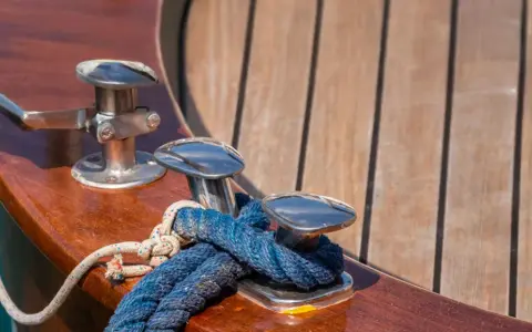 Corde bleue et blanche nouée sur des taquets chromés, prêts pour nettoyer le pont du bateau. Le bois verni brille au soleil.