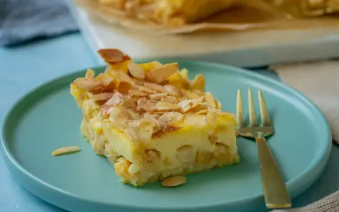 Ein Stück schneller Apfelkuchen mit Schmand und Pudding, bestreut mit Mandelblättchen und Zimt, auf einem türkisfarbenen Teller.