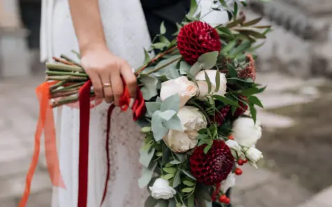 Dłoń panny młodej trzyma bukiet z bordowych dalii i kremowych róż, ozdobiony czerwonymi wstążkami. Idealne czerwcowe kwiaty na ślub.